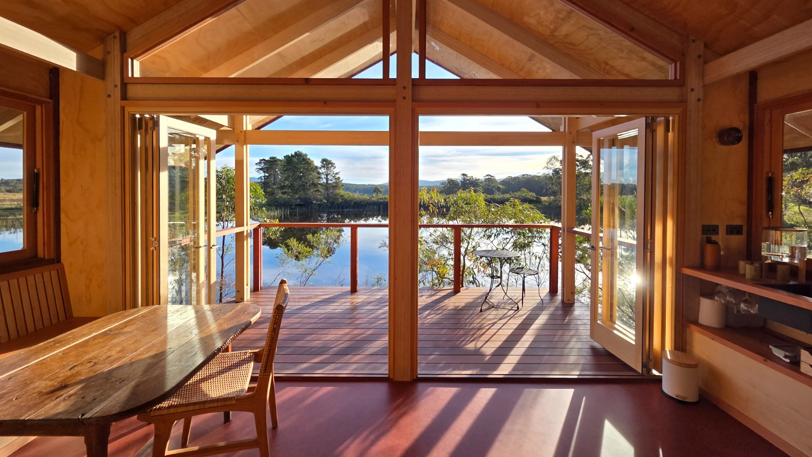 A timber and bamboo-lined interior with a pitched ply ceiling, opening through full-height glazed doors onto a deck above still water and bushland