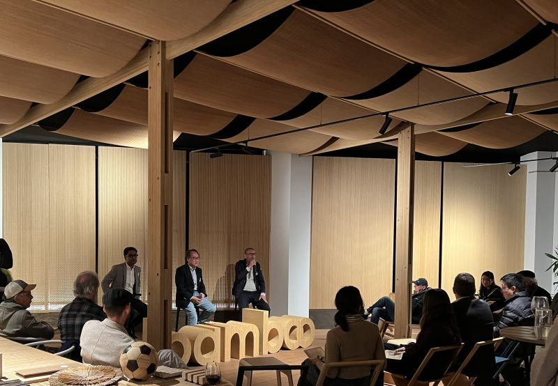An audience seated in a bamboo-lined space watching a panel discussion, with a large bamboo sign at the front of the room